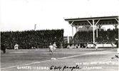 Original title:  Courtesy of Saskatoon Public Library. May 14, 1914. Postcard of Saskatoon's professional team, the Quakers, playing baseball against Regina in Saskatoon's new ball park, Cairn's field. The grandstand can be seen in background with 6404 people in attendance.