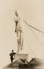 Original title:  Photographie d’archives en noir et blanc. Un homme est accoudé au pied d’une statue faisant plusieurs fois sa hauteur. La sculpture blanche représente la Vierge Marie couronnée d’étoiles, les mains jointes devant elle. La statue est posée sur un socle qui est lui-même placé sur le roc.