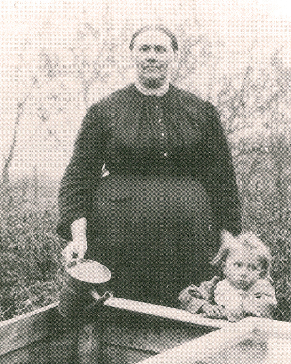 Titre original :  Anna Toews (1868-1933) with her daughter Margaret (1908-25) standing behind a cold frame which was used to start plants. Taken around 1912/13 which would have been shortly after they returned from Needles, BC.
Source: Cathy Barkman, “Anna Toews (1868–1933): midwife,” Preservings (Steinbach, Man.), 10 (June 1997), pt.2: 51. [https://www.plettfoundation.org/files/preservings/Preservings10-2.pdf] 